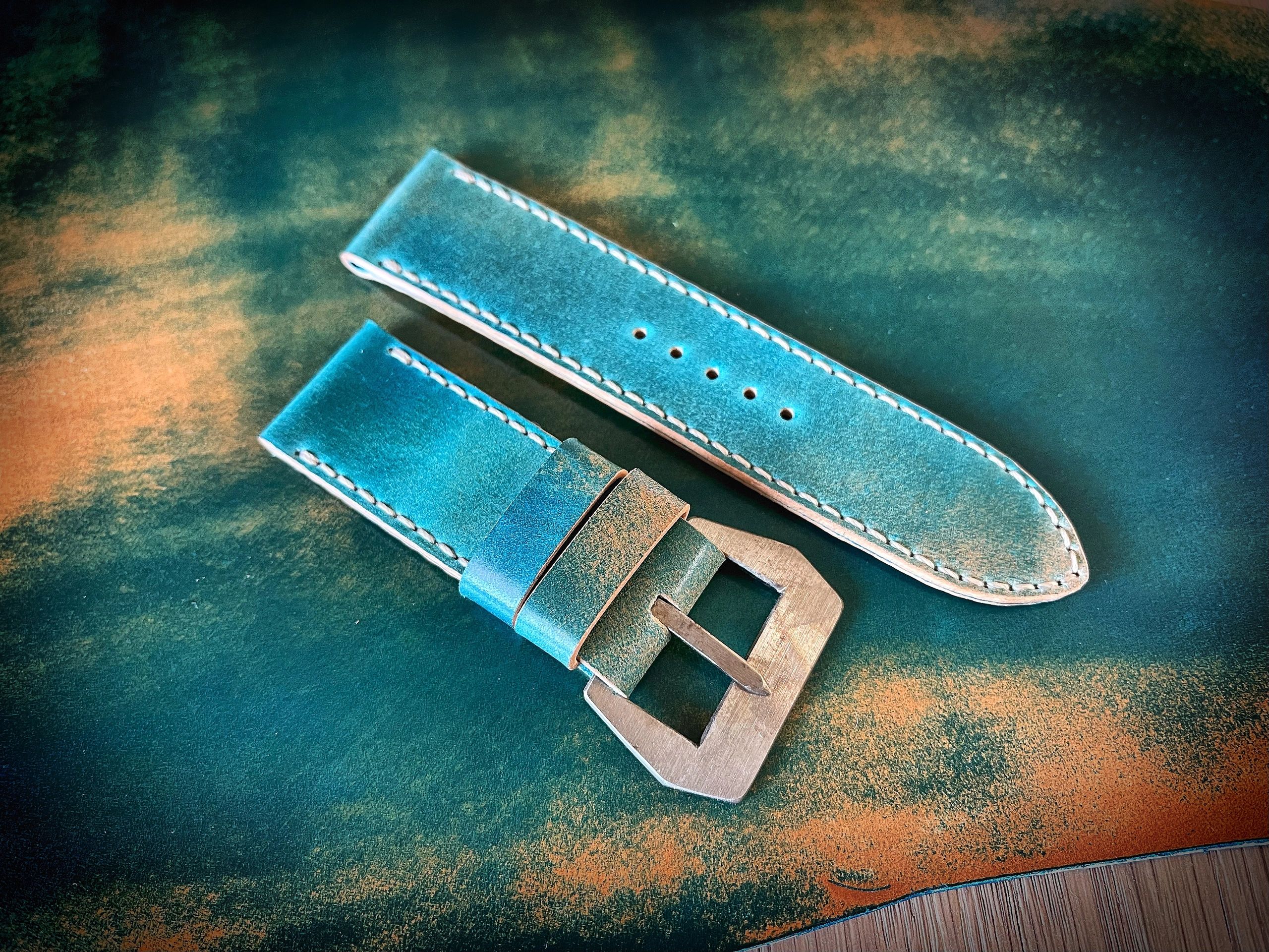 Handcrafted teal leather watch strap with silver buckle on a textured surface.