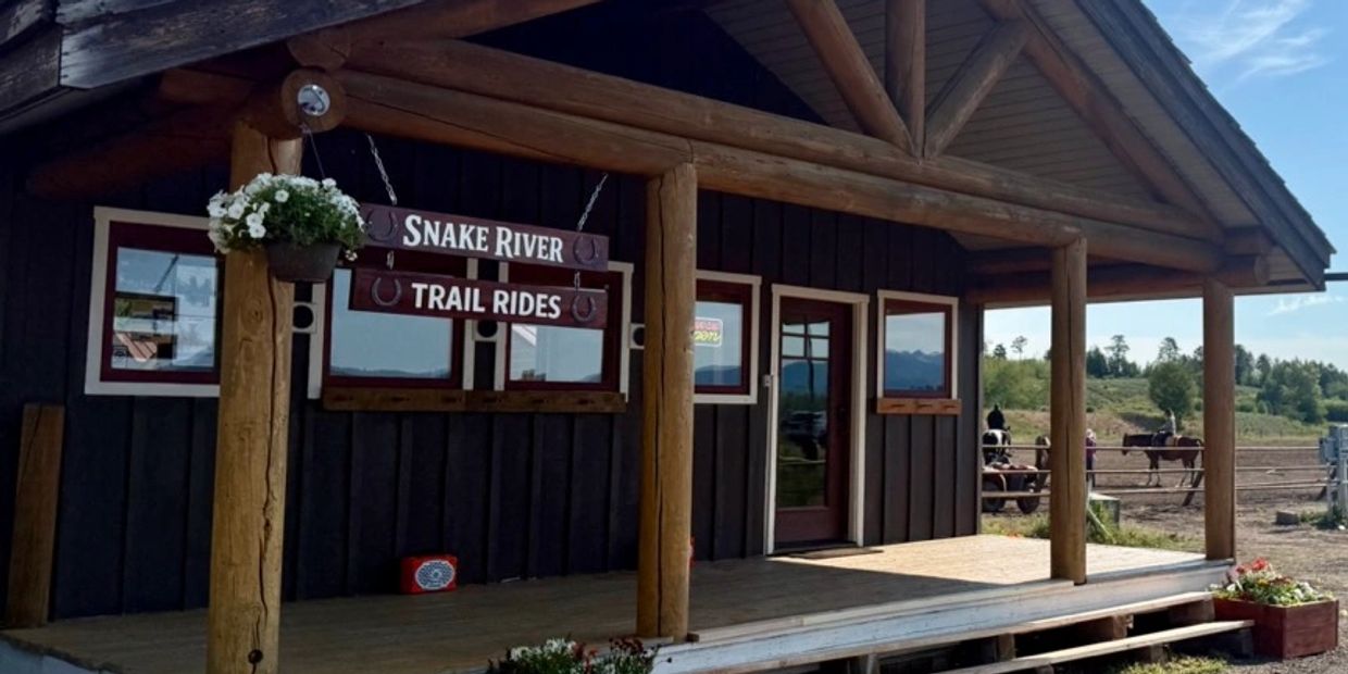 Rustic building with Snake River Trail Rides sign and horses in the background.