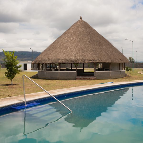 Thatched roof hut near a calm swimming pool under a cloudy sky.