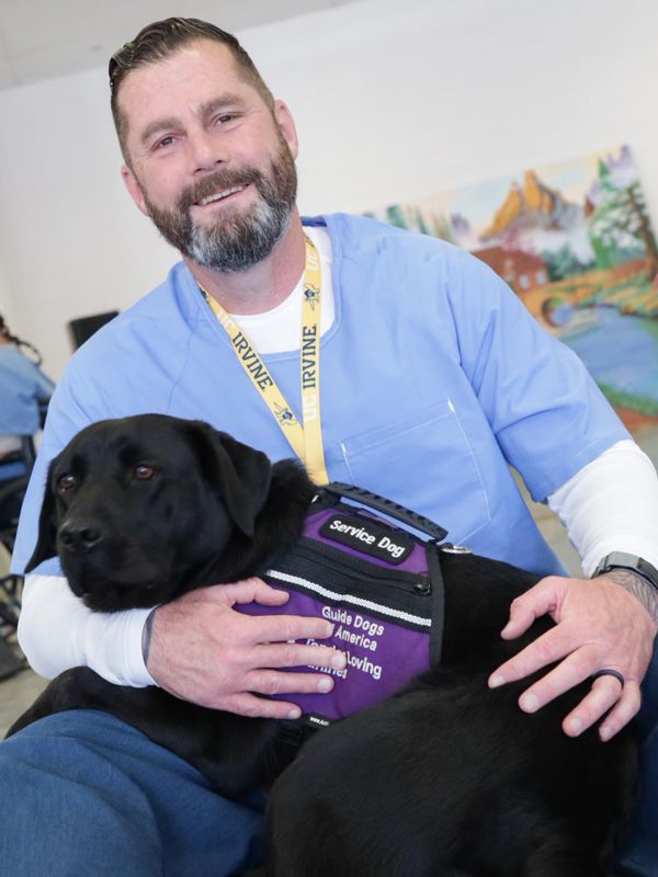 Man in blue scrubs with a black service dog wearing a purple vest.
