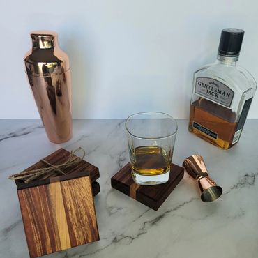 Whiskey setup with a glass, bottle, shaker, and wooden coasters on a marble surface.