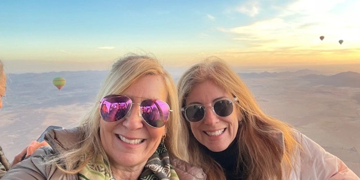Two women smiling in the foreground, both wearing sunglasses and jackets, with colorful hot air ball
