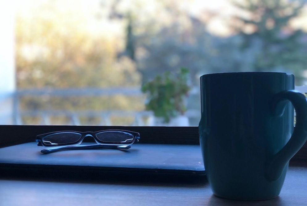 A laptop computer, pair of reading glasses and coffee mug sit on a desk.