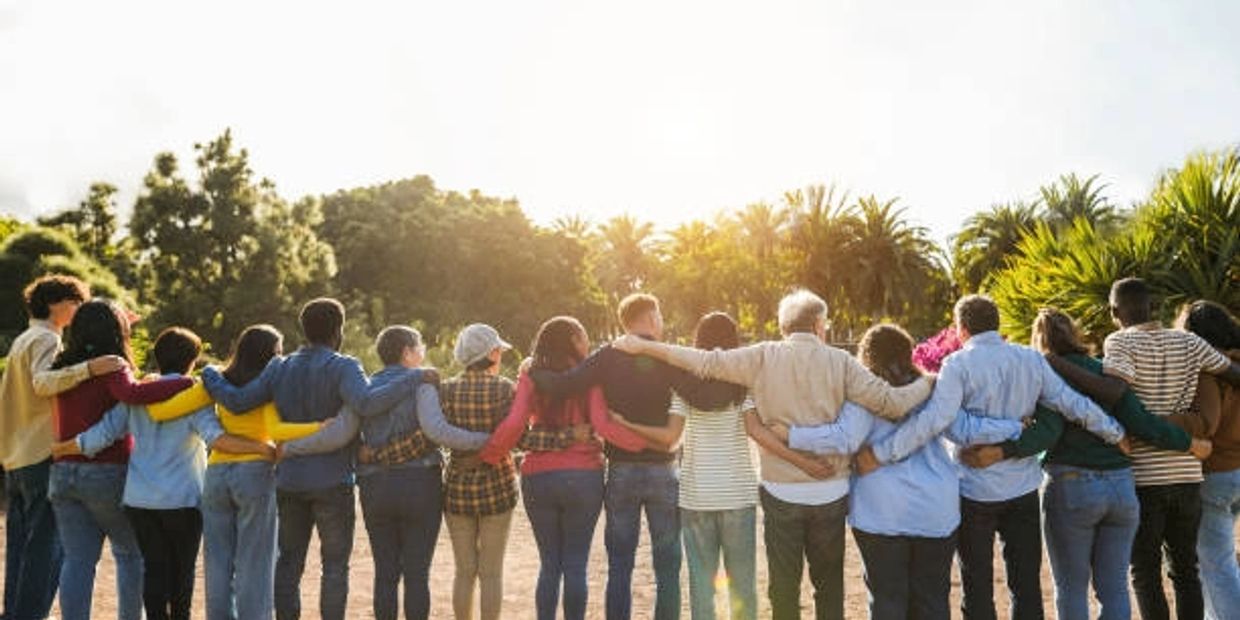 A diverse group standing arm in arm outdoors with sunlight and trees.