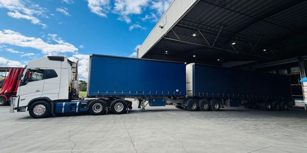 A large white truck with two blue trailers parked outside a warehouse under a blue sky.