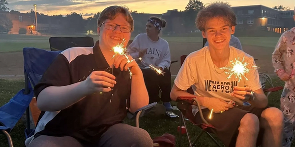Two young men smiling while holding sparklers at dusk.