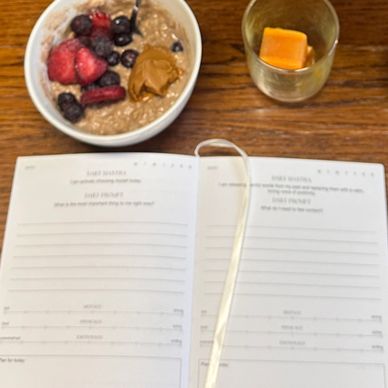 A breakfast setup with flowers, oatmeal, candle, vitamins, and an open planner on a wooden table.
