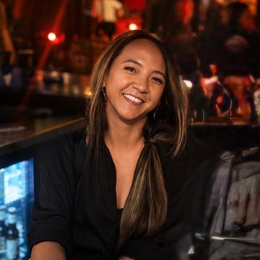 Smiling woman with long hair in a cozy, dimly lit bar setting.