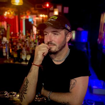 Man with tattoos and a cap sitting thoughtfully in a dimly lit bar.
