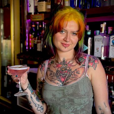A colorful-haired woman with tattoos holds a cocktail in a bar setting.