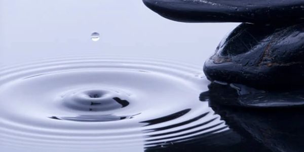 A water droplet creating ripples near smooth black stones in calm water.