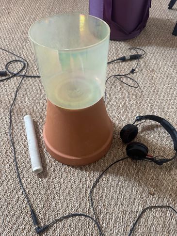 A terracotta pot with a translucent plastic container on top, surrounded by headphones and cables on carpet.