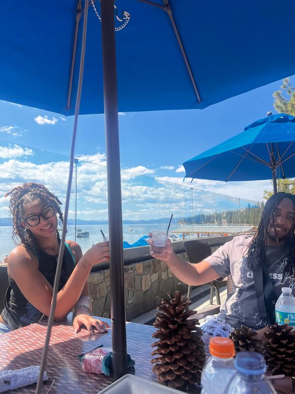 Two women enjoying drinks outside under a blue sky and umbrella.