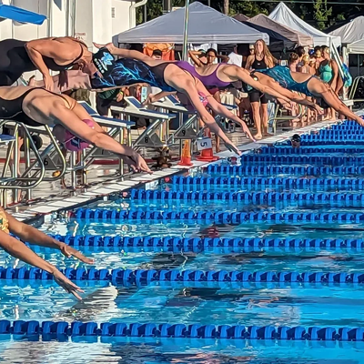 Swimmers diving into the pool at the start of a race in a sunny outdoor competition.