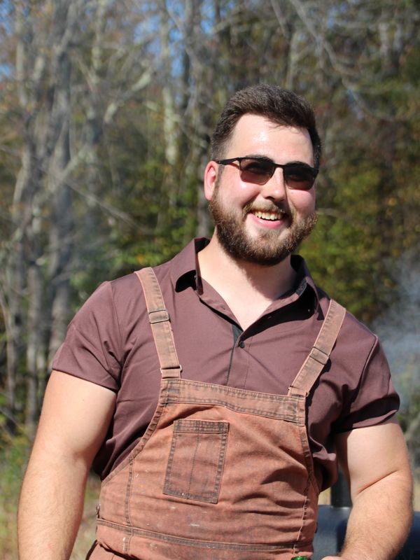 A man wearing sunglasses and brown overalls smiling outdoors.