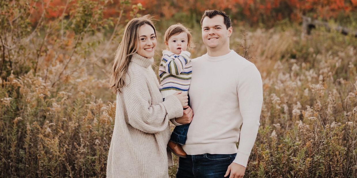 A smiling family of three stands outdoors in autumn.