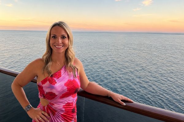Woman in floral dress smiles on a balcony overlooking the ocean at sunset.