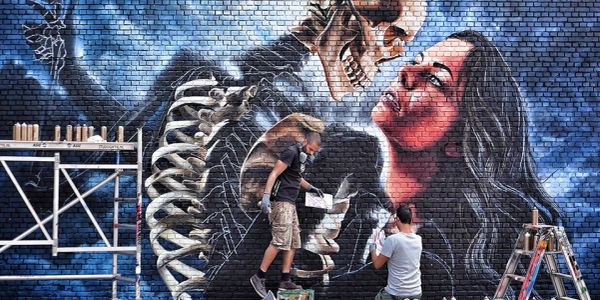 Artists painting a dramatic mural of a skeleton and woman on brick wall.