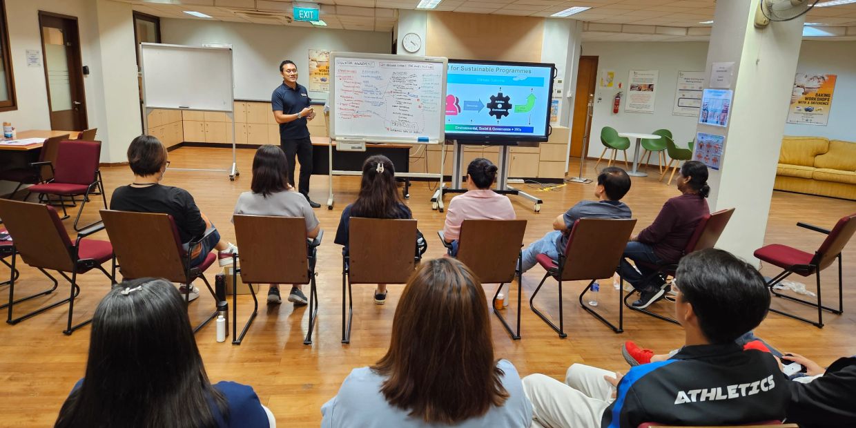A man presents to a seated group in a classroom with whiteboards and a screen.