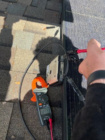 Person testing a solar panel with a digital clamp meter on a rooftop.