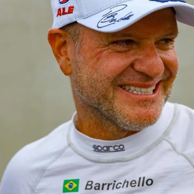 Smiling man in racing gear with Barrichello name and Brazil flag.