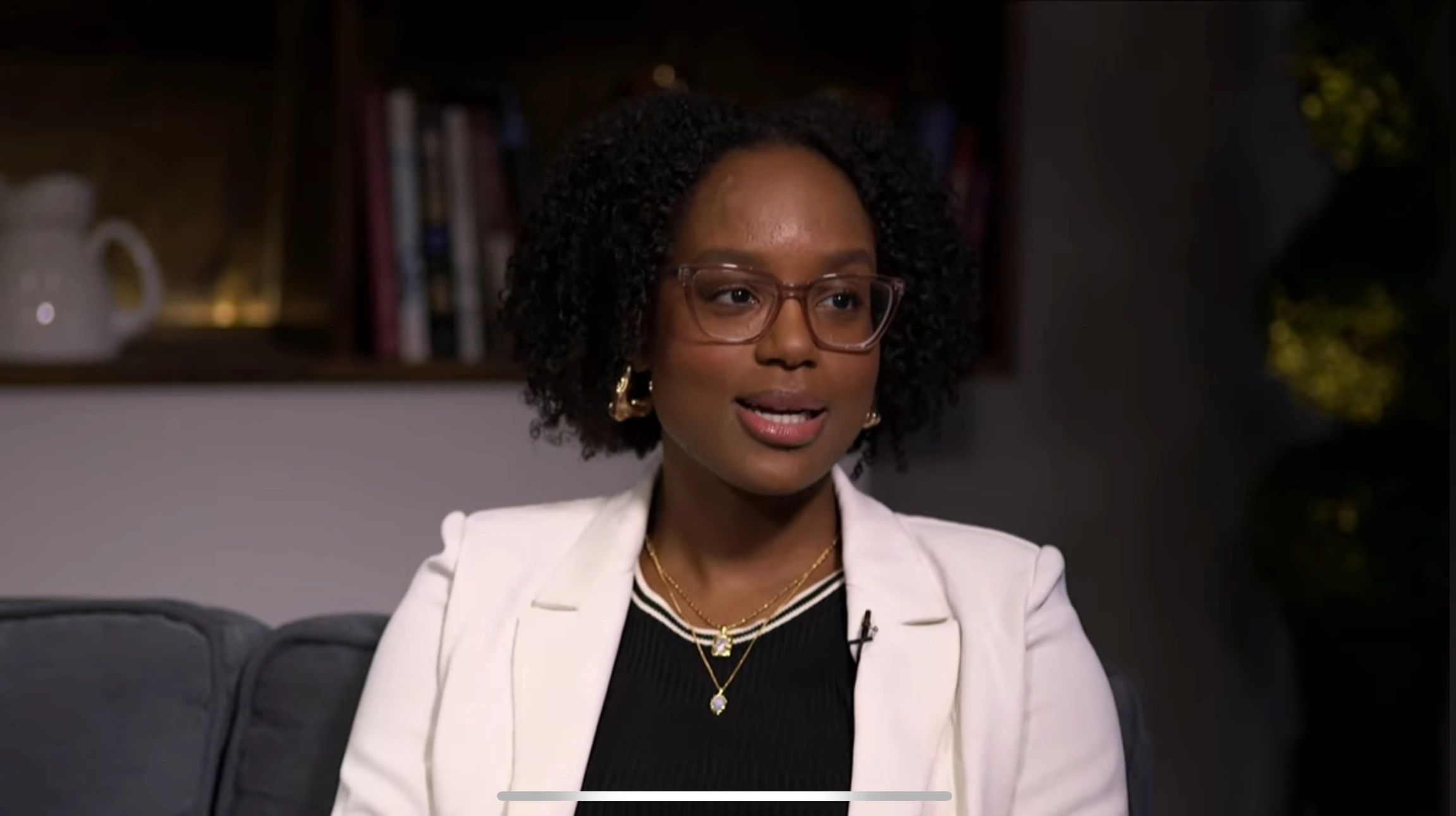 Woman with curly hair and glasses speaking in a cozy setting.