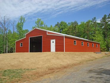 Large red metal barn on a dirt hill with a forest backdrop.