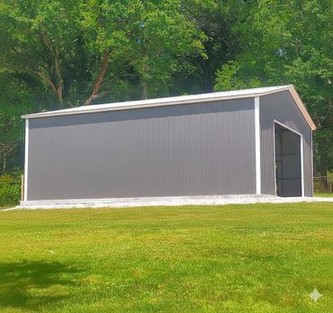 A large gray metal storage shed on a concrete slab surrounded by green grass and trees.