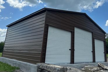 Large brown metal building with three white roll-up doors under a partly cloudy sky.