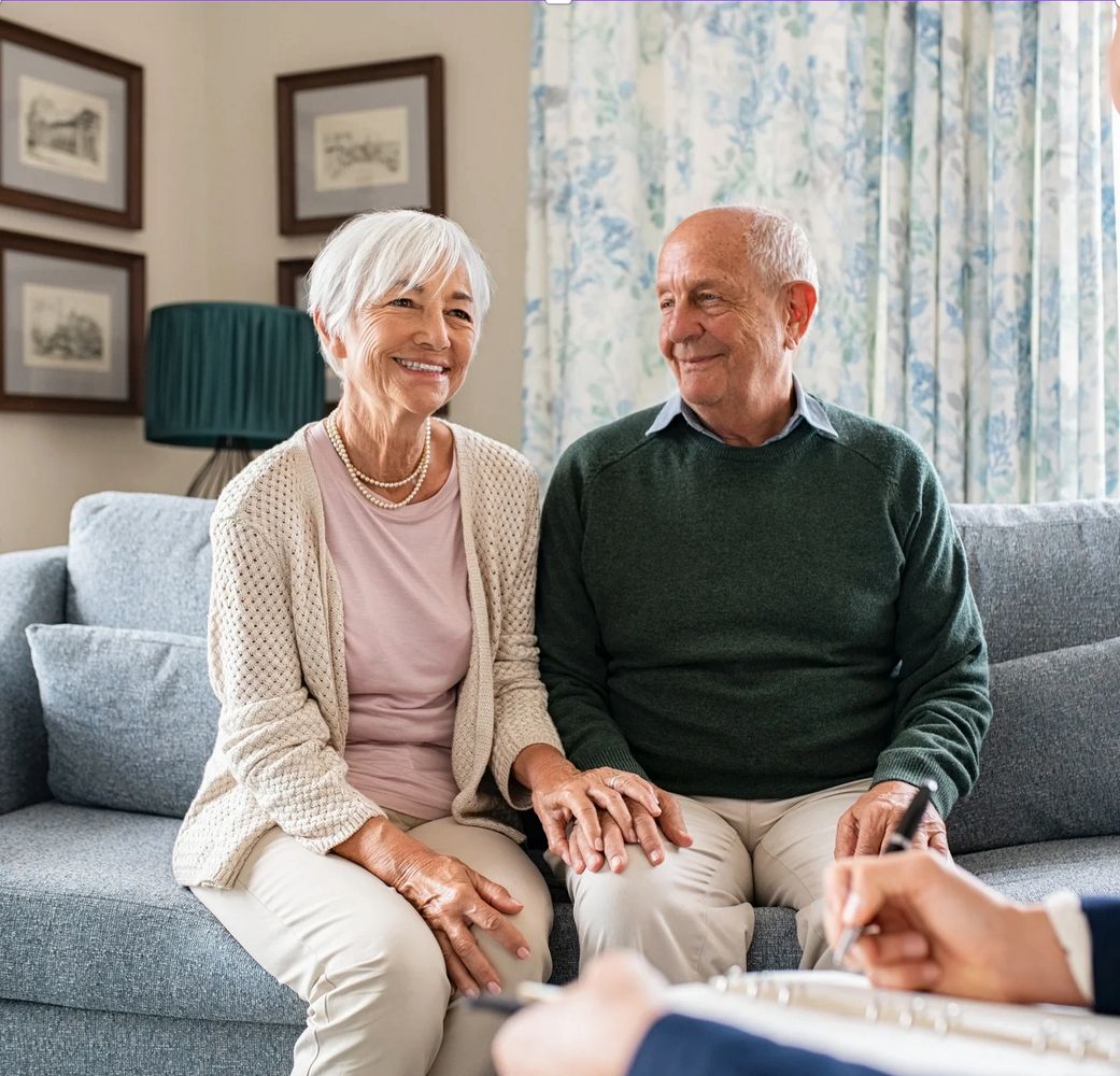 Elderly couple sitting on a couch, holding hands and smiling during a consultation.