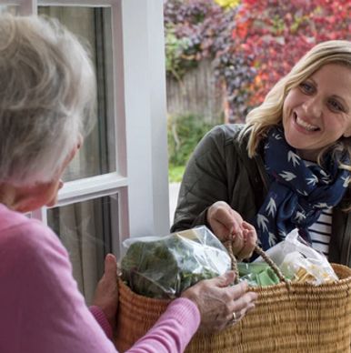 A young woman hands a basket of groceries to an elderly woman at the door.