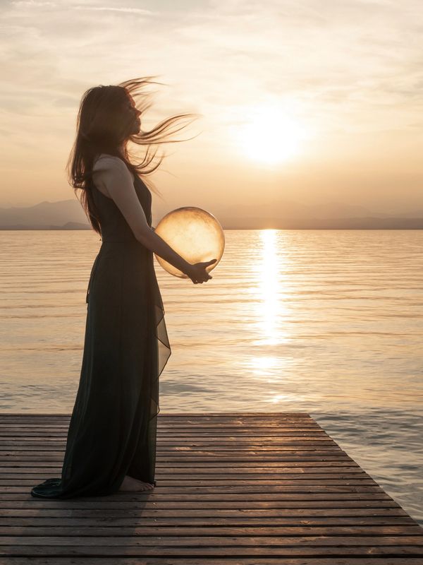 Woman holding a glowing orb by the water at sunset.