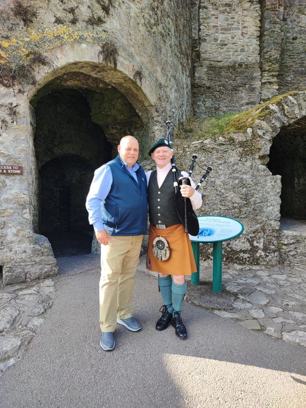 Two men stand in front of a stone archway, one holding bagpipes and wearing traditional Scottish attire.