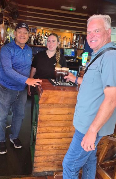 Three people at a bar, two men and a bartender with drinks.