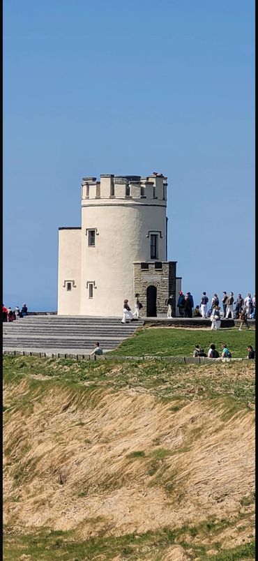 Historic stone tower on a coastal cliff with visitors around it.