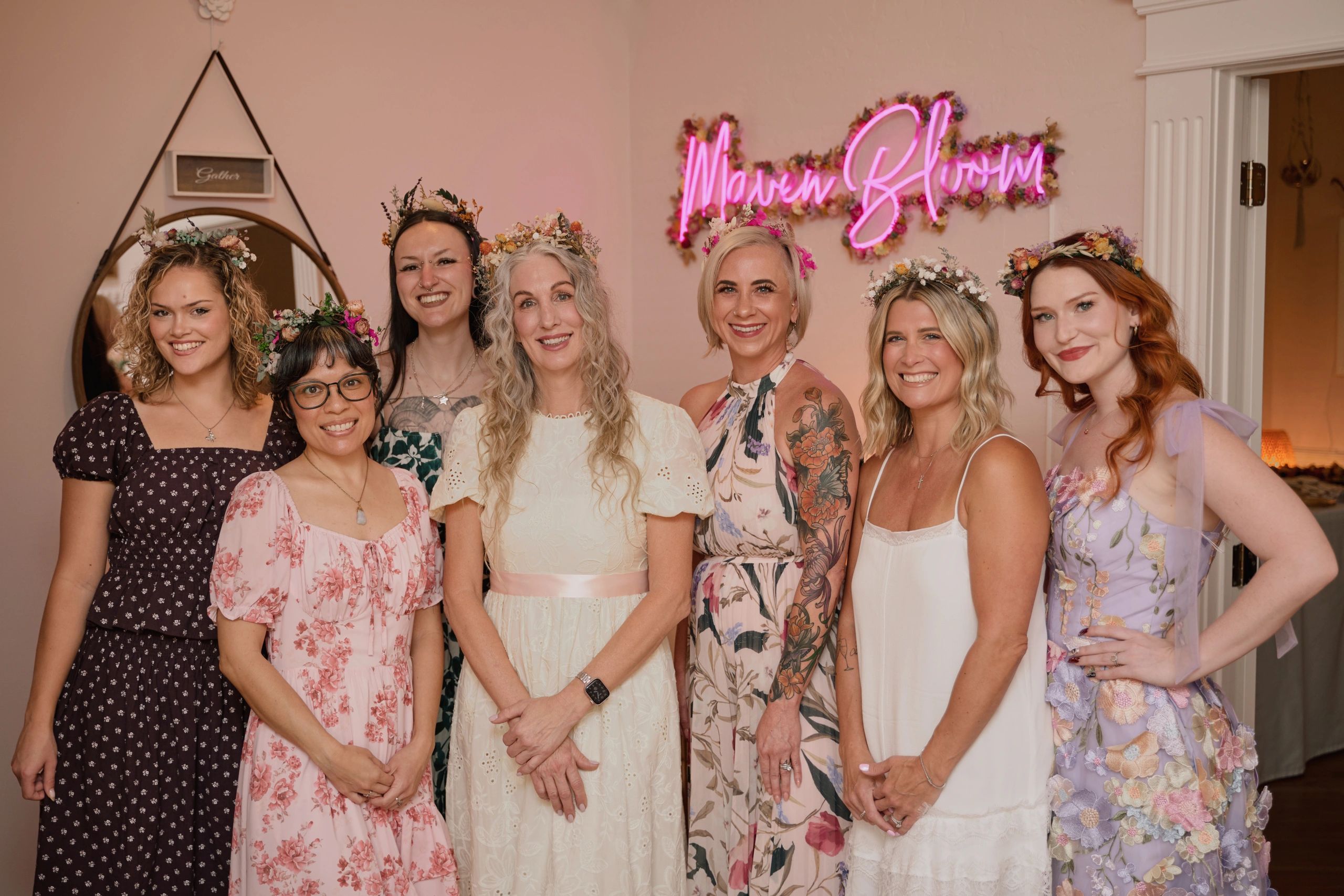Seven women wearing floral crown headpieces smiling indoors.
