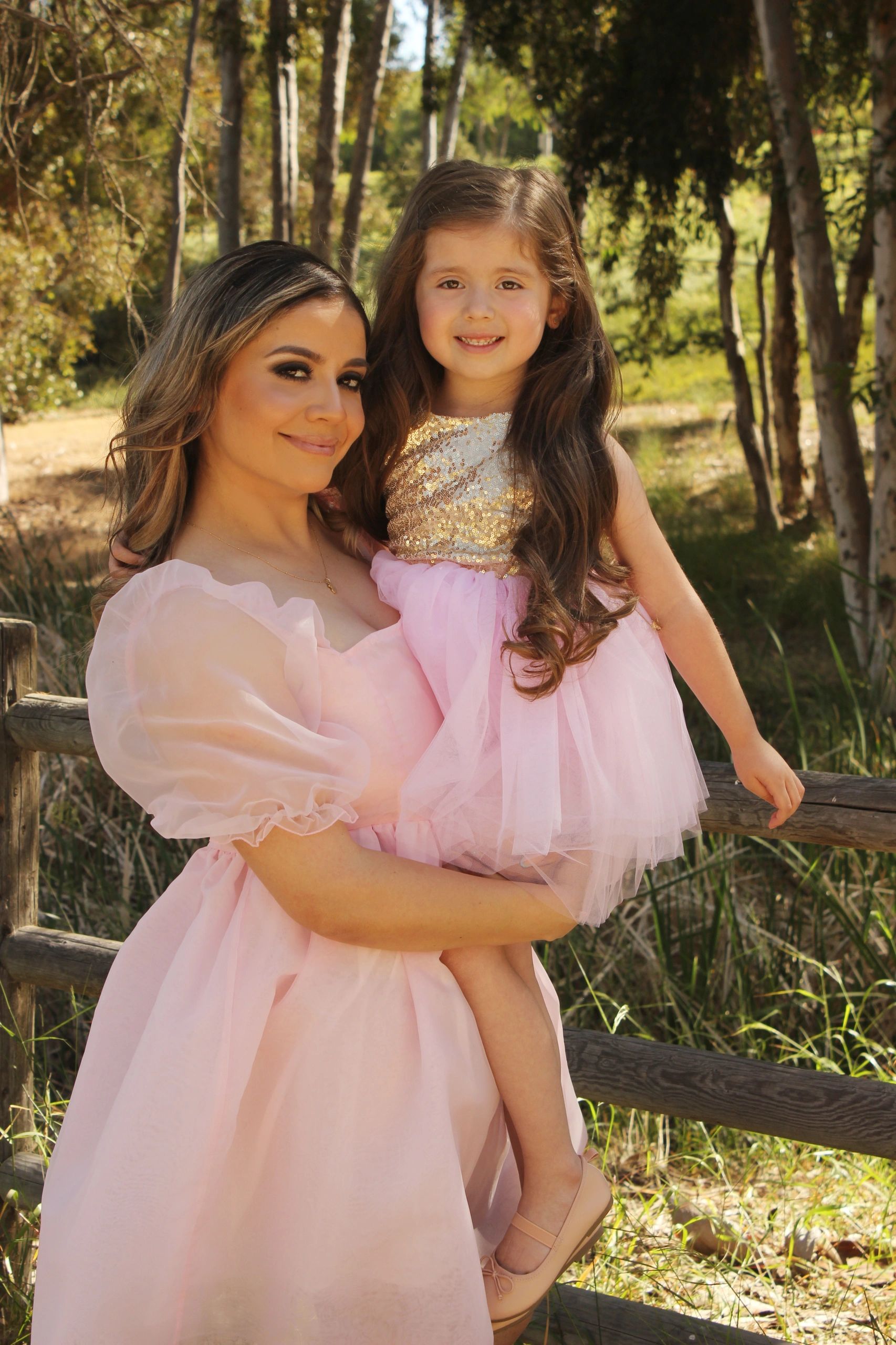 A woman holding a young girl, both in pink dresses, outdoors by a wooden fence.