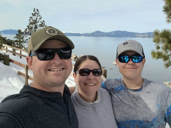 Three smiling people wearing sunglasses and hats by a snowy lakeside with mountains in the background.