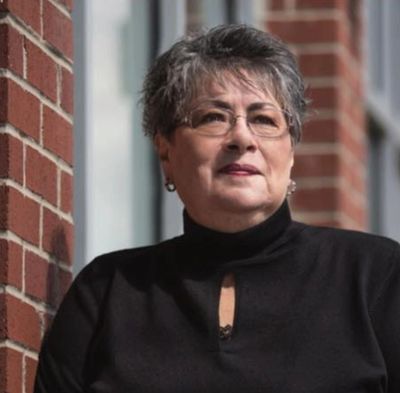 Older woman with glasses leaning against a brick wall, wearing a black top.