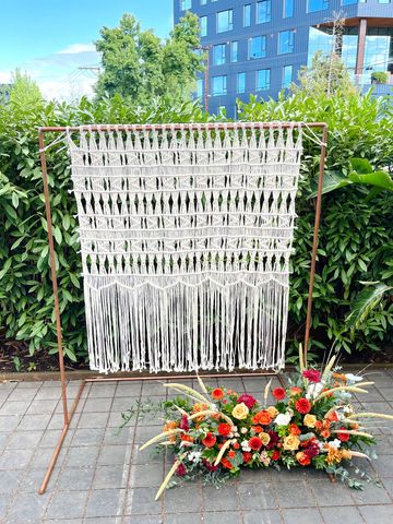 Copper pipe ceremony backdrop frame with macrame hanging and floral arrangement in the foreground.