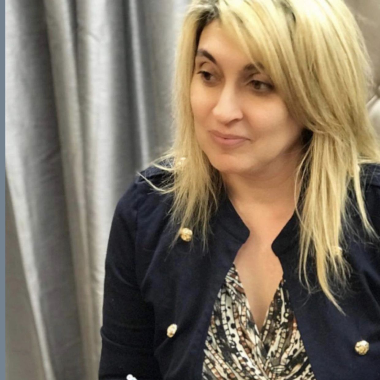 A woman with blonde hair wearing a patterned top and dark jacket.