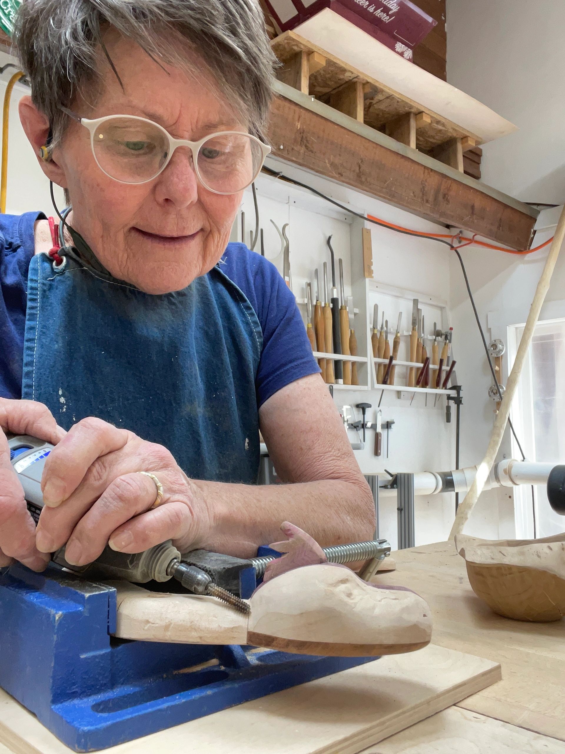 Elderly woman carving wood in a workshop with tools on the wall.