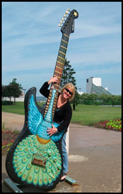 Woman posing with a giant colorful guitar sculpture outdoors.