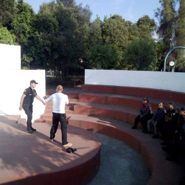 A police officer training a man in front of seated individuals outdoors.