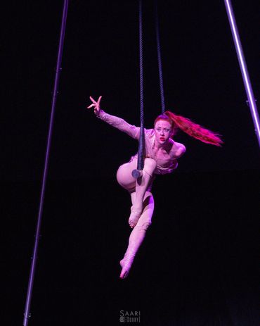 Aerial performer suspended mid-air with bright red hair against a dark background.