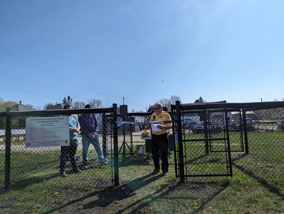 Man speaks at the entrance of a fenced dog park on a sunny day.