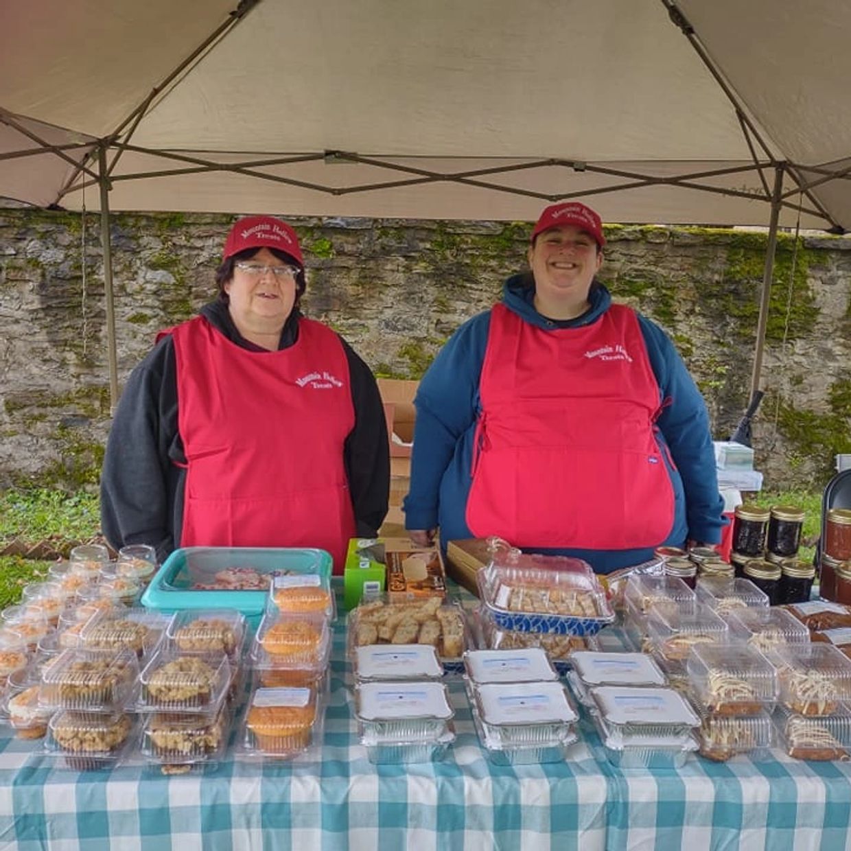 Two women in red aprons and hats selling baked goods and jams at a market stall.