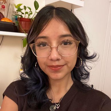 Young woman with glasses and curly dark hair smiling indoors.