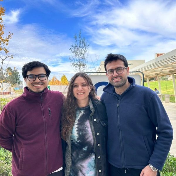 Three friends smiling outdoors under a partly cloudy sky.