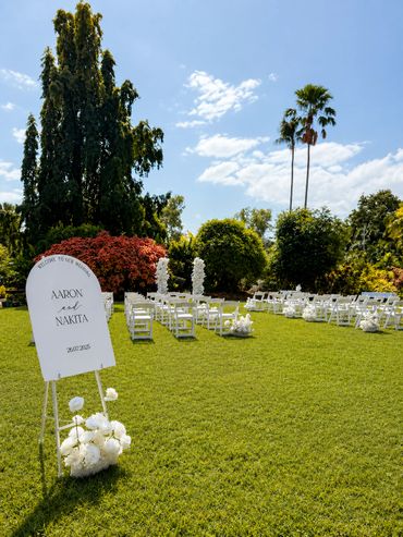 Outdoor wedding setup with white chairs and floral decorations on a sunny day.